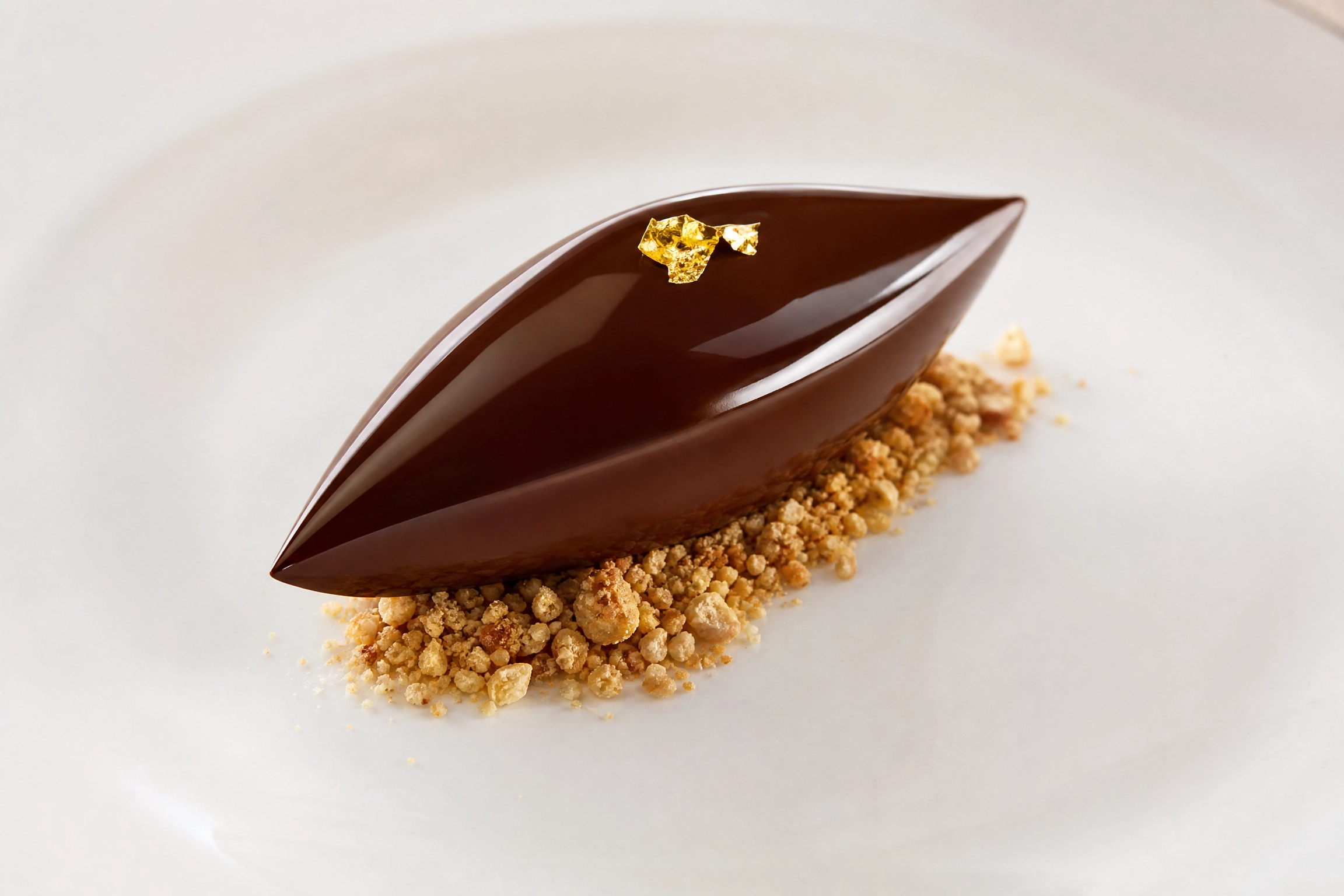 Glossy dark chocolate quenelle with gold leaf accent on a hazelnut praline crumble base